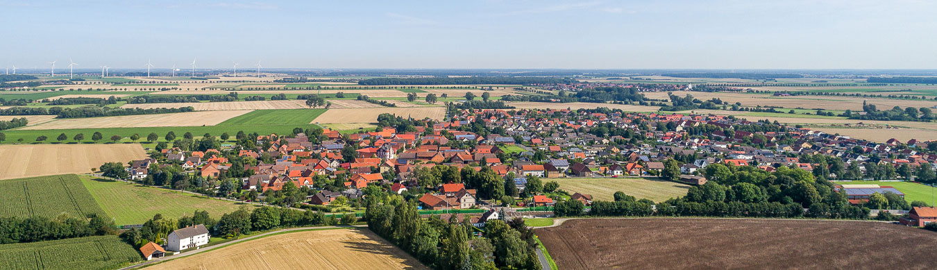 Ein Dorf aus der Vogelperspektive. Es ist freundliches sonniges Wetter. Am Horizont ist ein blauer fast wolkenloser Himmel. Die Häuser des Dorfes haben fast alle rote Dächer. Im Vordergrund sind zwei Felder. Im Hintergrund sind viele landwirtschaftlich genutzte Flächen und kleinere Wälder. Links am Horizont stehen einige Windkrafträder.
