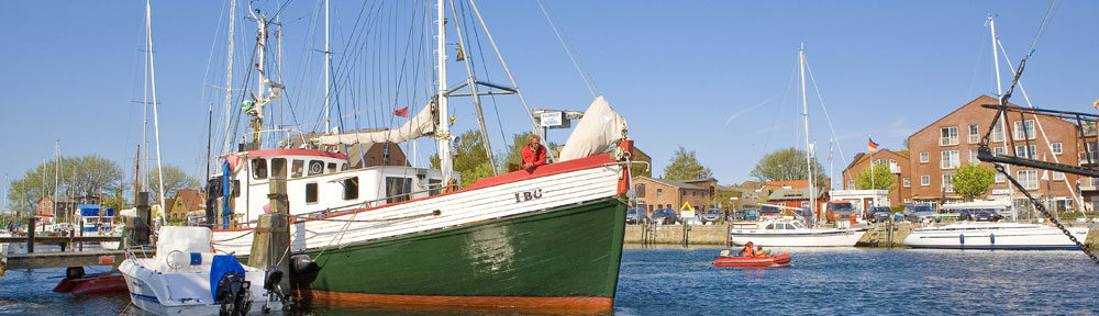 Auf dem Bild ist ein grün-weißes Segelschiff zu sehen. Das Schiff ist ca. 20 Meter lang. Es nimmt fast die Hälfte des Bildes ein. Das Schiff liegt in einem Ostsee-Hafen vor Anker. Es ist sonniges Wetter, das Wasser ist blau und der Himmel wolkenlos. Im Hintergrund ist ein Hafenort zu erkennen. Die Häuser bestehen aus rotem Ziegelstein. Auch andere Schiffe und Boote sind auf dem Wasser. Auf dem Schiff steht am Bug ein Mann mit roter Jacke und telefoniert mit dem Handy und schaut dabei aufs Wasser.