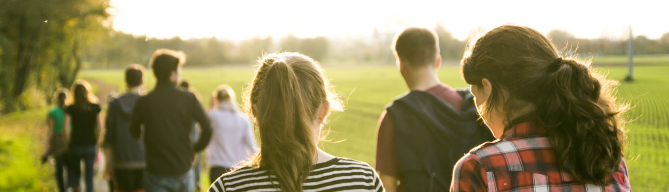 Eine Gruppe von Jugendlichen geht dem Sonnenuntergang entgegen. Sie sind nur von hinten zu sehen. Im Vordergrund sind zwei Mädchen. Die rechte hat längere dunkle Haare, die linke hat blondes Haar. Vor ihnen gehen ca. 8 andere Jugendliche. Sie gehen auf einem Feldweg. Rechts neben ihnen ist ein grünes Feld, links beginnt ein Wald.
