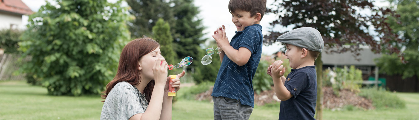 Eine junge Frau pustet Seifenblasen Richtung zweier kleiner Jungen. Die drei sind in einem sommerlichen Garten auf einer größeren Wiese. Die Betreuerin sitzt links und die beiden Jungen stehen rechts daneben. Sie sind auf Augenhöhe. Man sieht die drei von der Seite. Der Junge in der Mitte ist ca. 5 Jahre alt. Er kneift die Augen zu und hält schützend seine Arme gegen die Seifenblasen hoch. Der rechte Junge, ca. 3 Jahre alt, trägt eine Schiebermütze und schaut auf die Seifenblasen und lächelt dabei.
