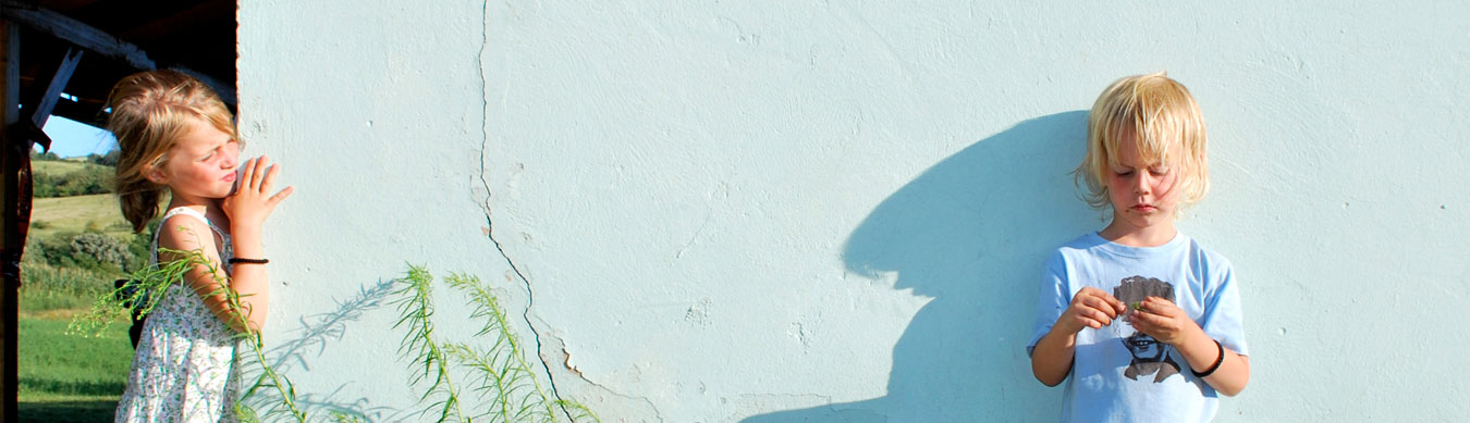 An einer weißen Hauswand steht ganz links ein blondes Mädchen mit einem hellen Sommerkleid. Ganz rechts im Bild steht ein kleiner blonder Junge mit einem hellblauen T-Shirt mit Aufdruck. Er wirft einen langen Schatten an die Wand. Beide sind ca. 4 bis 5 Jahre alt. Das Mädchen steht links an der Ecke der Hauswand und schaut auf den Jungen, der ein paar Meter entfernt steht. Der kleine Junge hält etwas Kleines in seinen Händen und schaut konzentriert darauf.